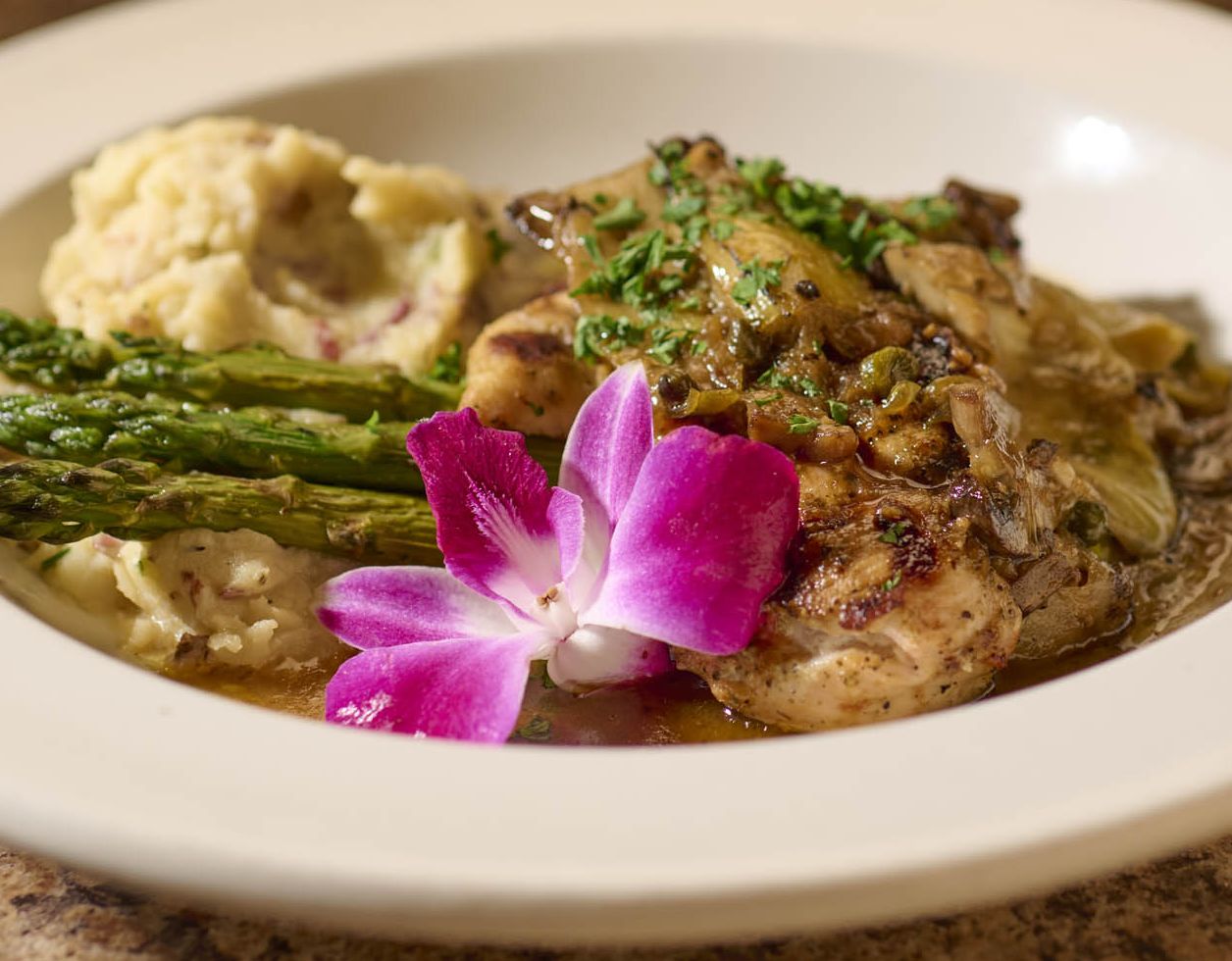 A plate of chicken marsala with asparagus and potatoes, with a colorful flower for garnish