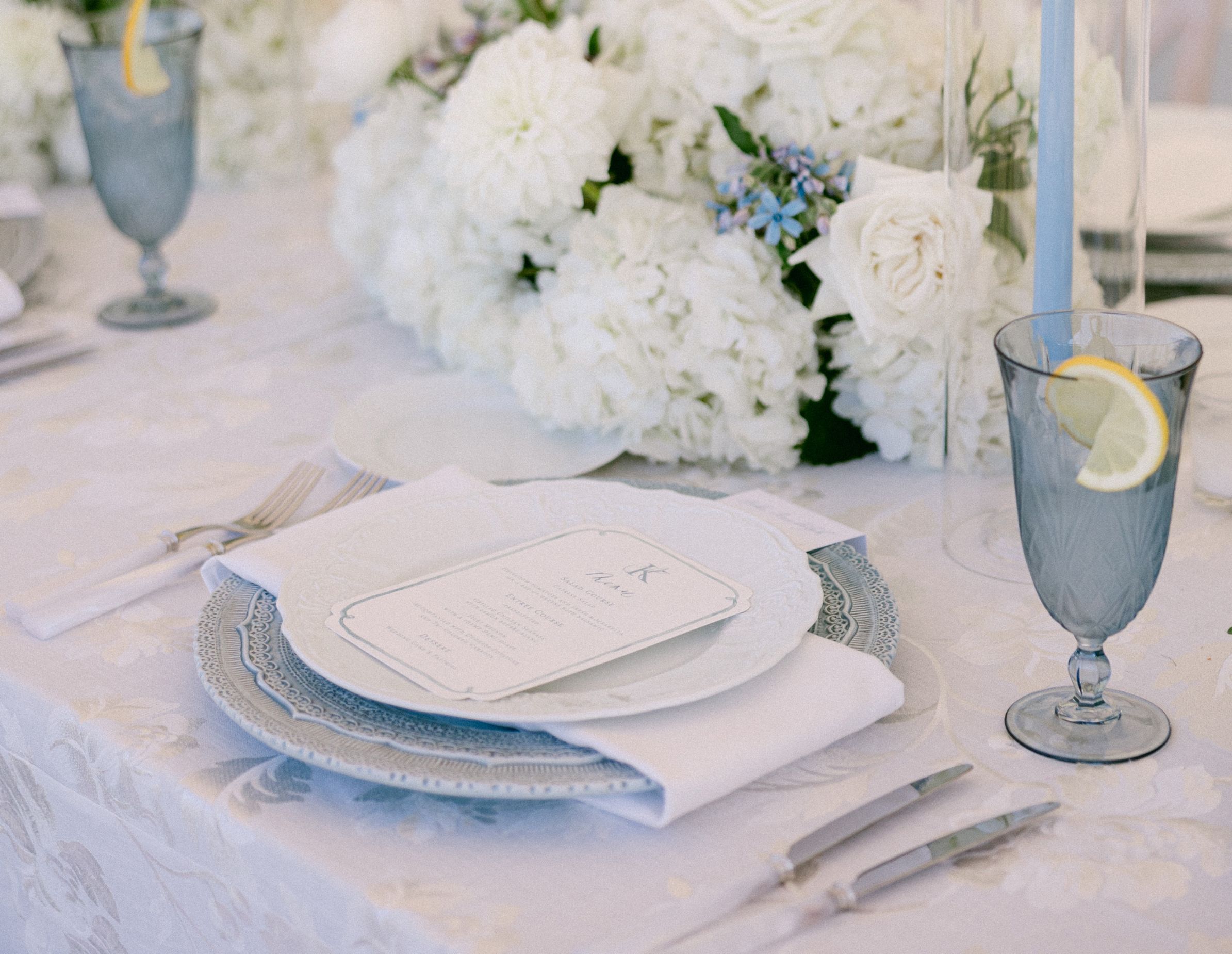 An elegant table setting featuring white china, a guest invitation, white flowers, and several wine glasses and flutes