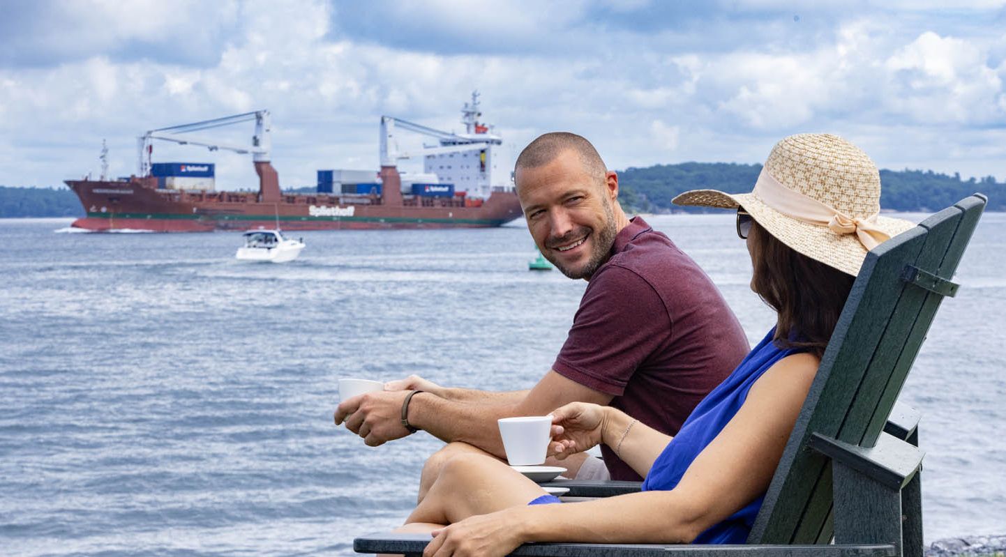 A couple relaxes in Adirondack chairs while a large steamer ship passes along the river in the background